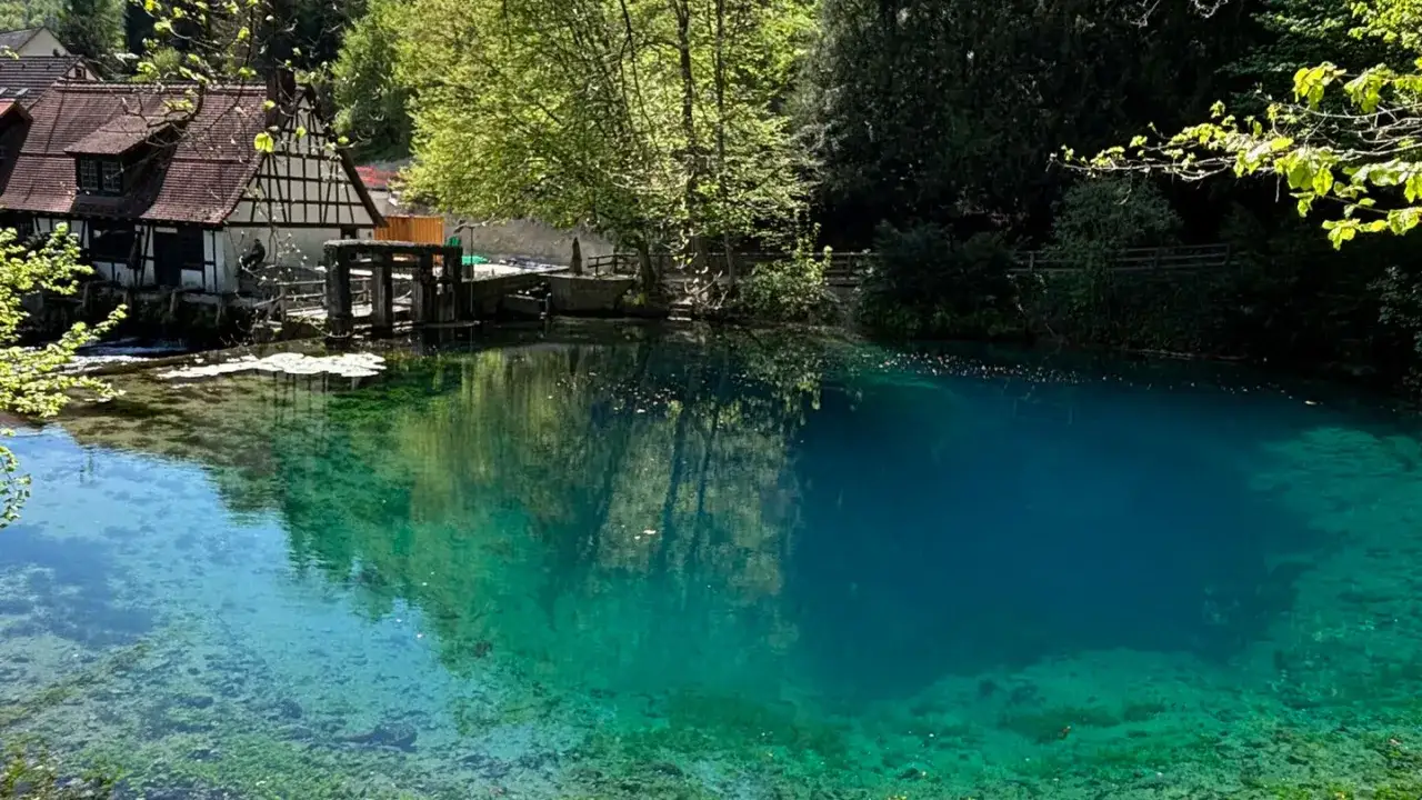 Der Blautopf See mit seinem klaren, blauen Wasser l&auml;dt zum Schwimmen ein. Ein historisches Geb&auml;ude am Ufer spiegelt sich im See.