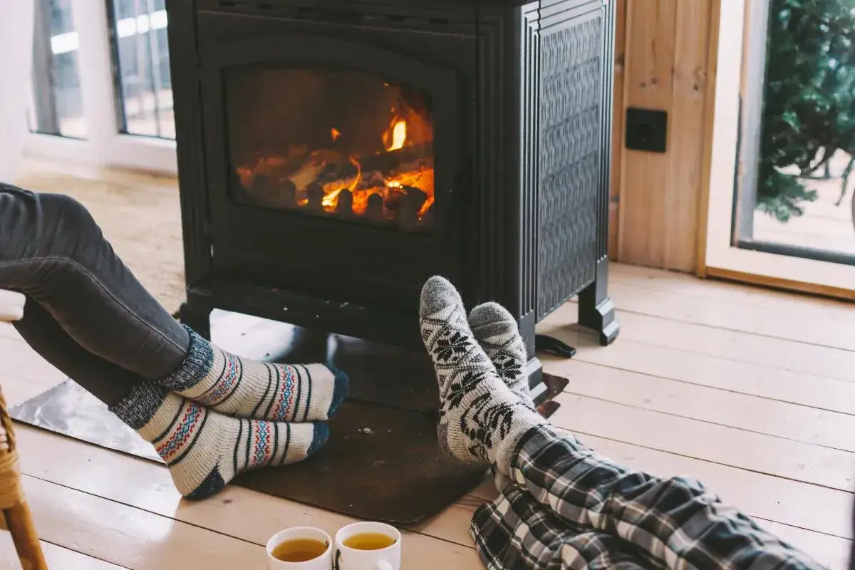 Pies abrigados con calcetines junto a un calefactor sin electricidad, disfrutando del calor de la chimenea y una taza de t&eacute;.