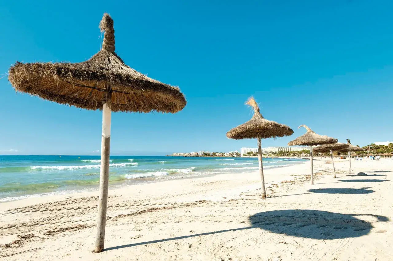 Soliga str&aring;hattparaplyer p&aring; en sandstrand vid Palma de Mallorca. V&aring;gor skvalpar in mot kusten under en klarbl&aring; himmel.