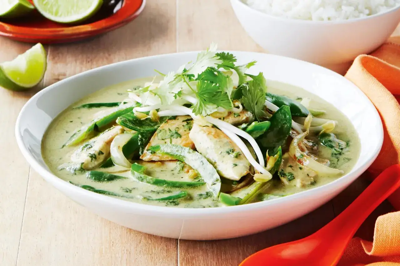 Pyszny green curry przepis z kurczakiem, zieloną papryką i kiełkami fasoli mung, udekorowany kolendrą. Obok miska ryżu i ćwiartki limonki.