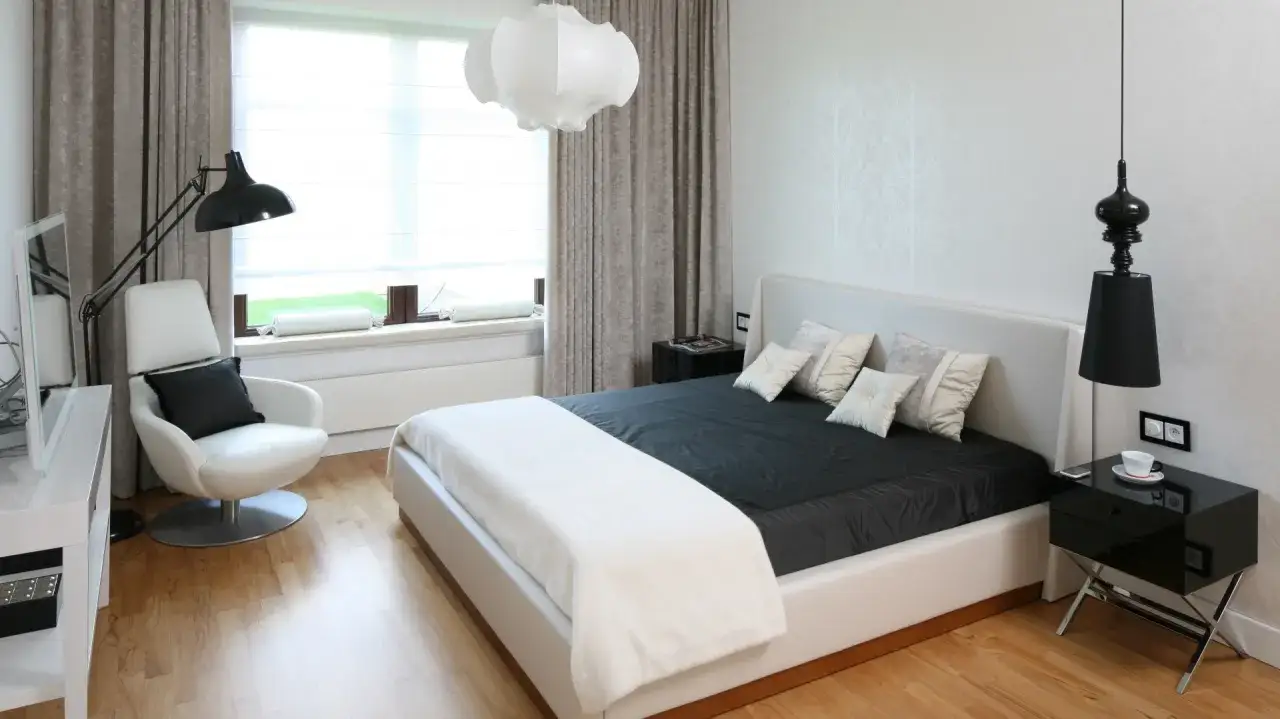 Nowoczesna sypialnia z białym ł&oacute;żkiem, czarną pościelą i designerskimi lampami. Okna z zasłonami. Warto sprawdzić wymiary okien typowych tabela.