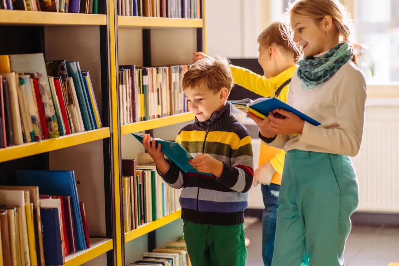 Dzieci w bibliotece wybierają lektury klasa 3. Chłopiec w kolorowym swetrze czyta książkę, dziewczynka z chustą na szyi przegląda kolejną.