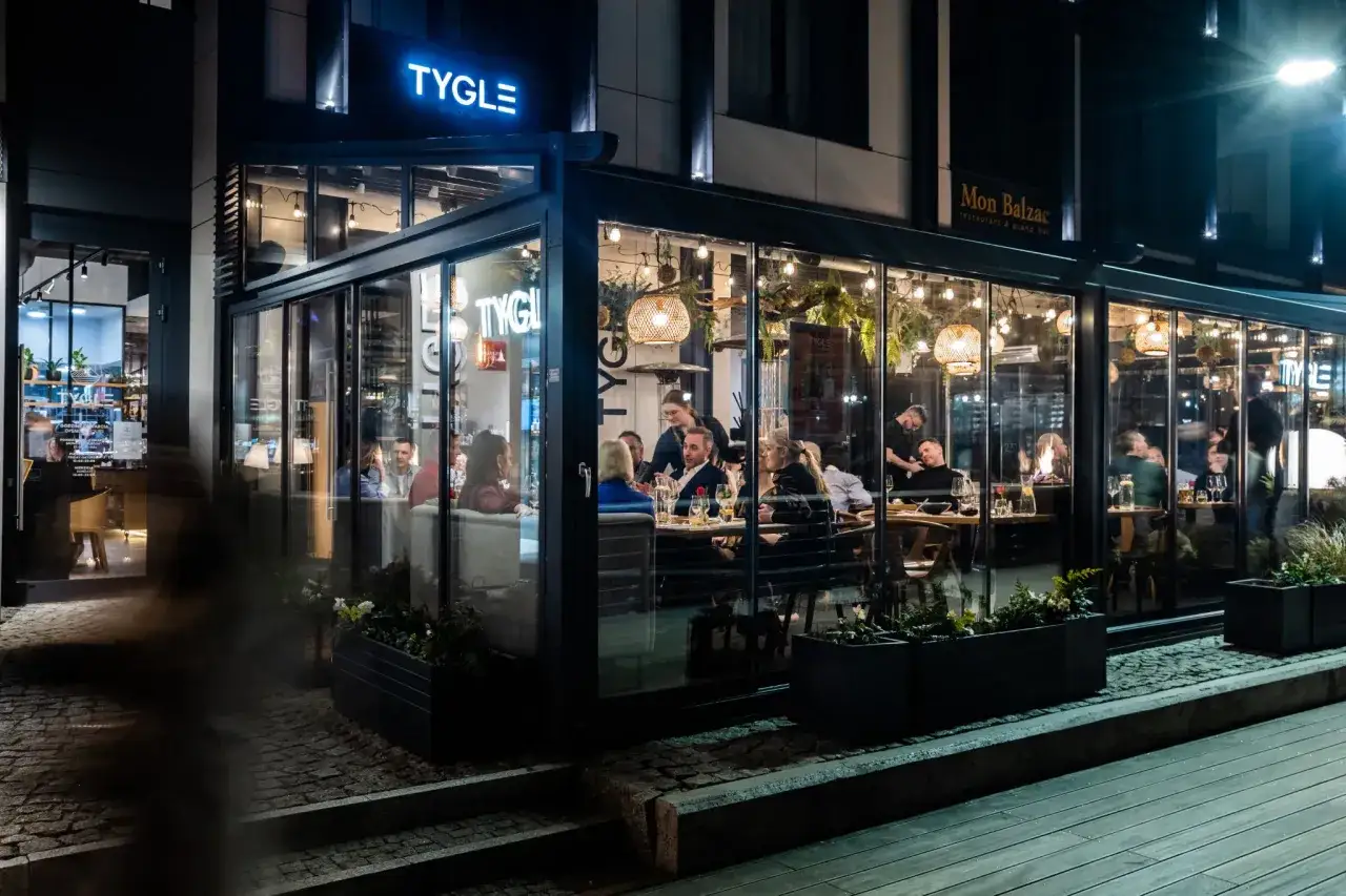 Wieczorem w restauracji Tygle Gdańsk przytulnie oświetlonej, ludzie siedzą przy stolikach, delektując się posiłkami i rozmowami.