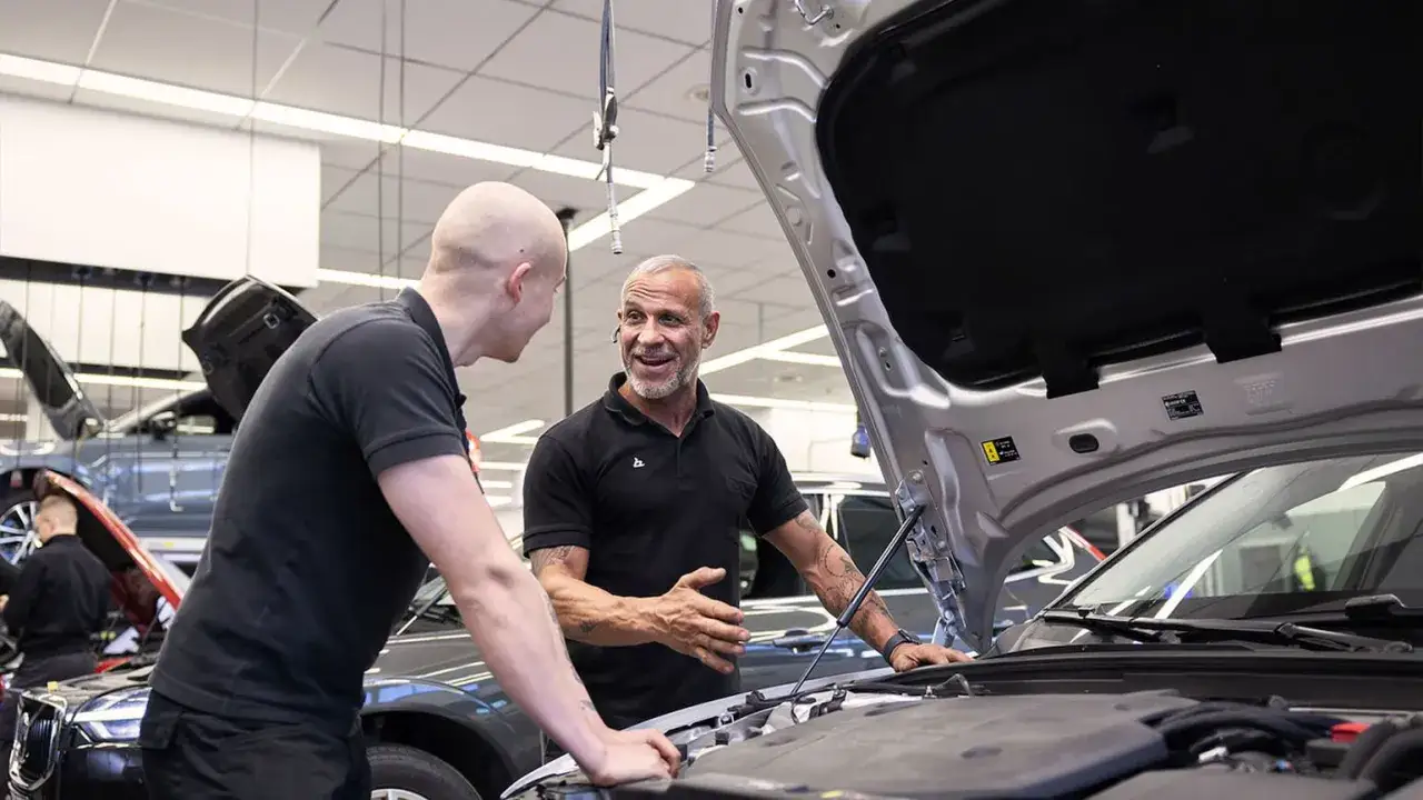 Två bilmekaniker diskuterar vid en öppen motorhuv. En av dem förklarar hur lång tid tar en bilservice, kanske för en kund.