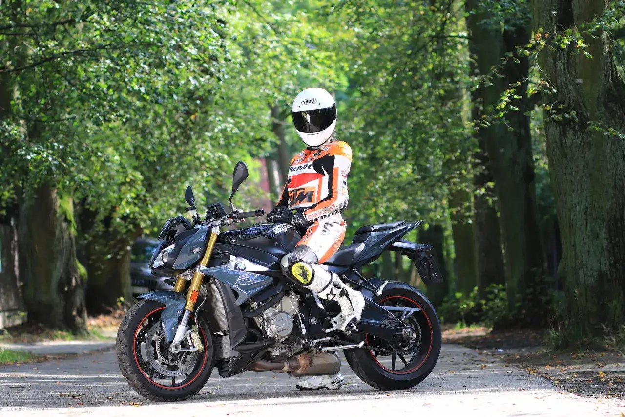 Motocyklista w kombinezonie siedzi na sportowym motocyklu BMW S1000R wśród drzew.
