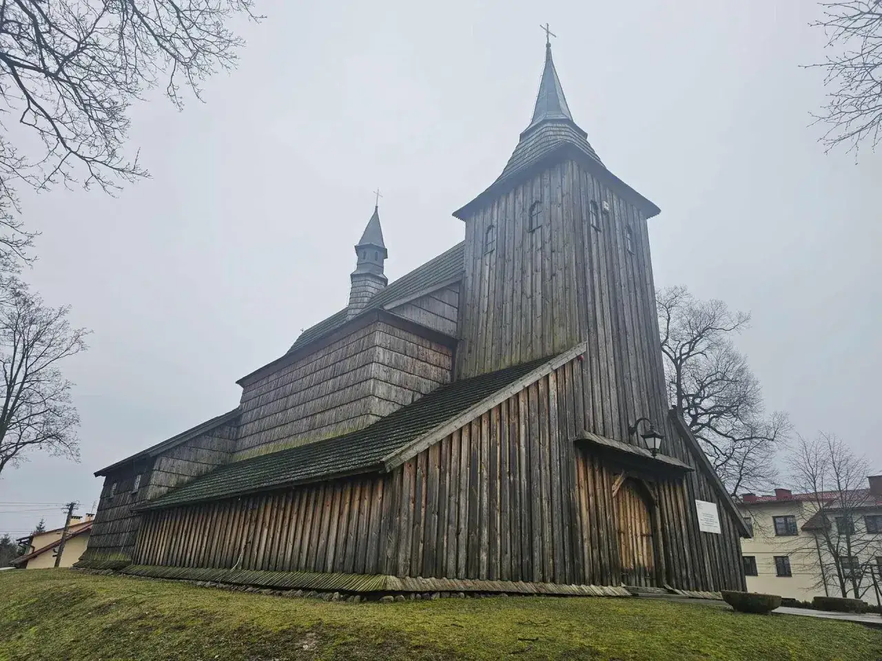 Drewniany kościół z wieżą i sygnaturką, pokryty gontem. W oddali widać budynki mieszkalne. To miejsce, gdzie czas się zatrzymał, jakby wyjęte z baśni o cmolas.