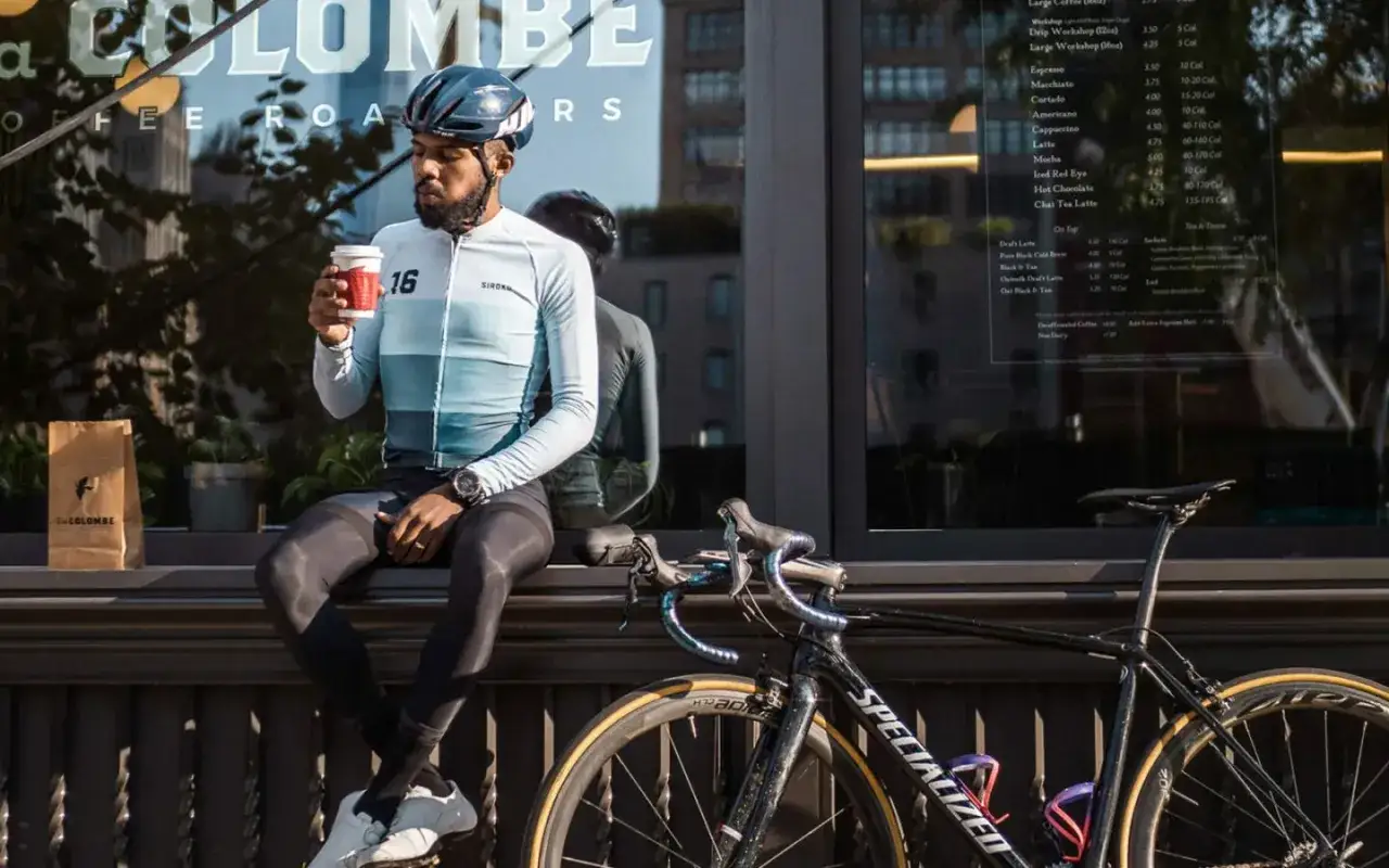 Kolarz w kasku i stroju kolarskim odpoczywa po coffee ride, pijąc kawę. Obok stoi rower.