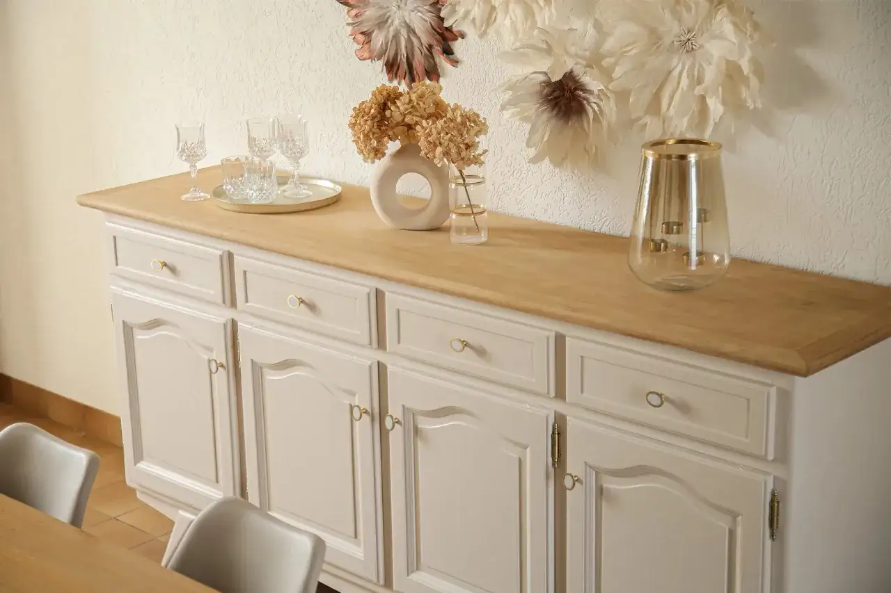 Un vieux buffet en bois relooké, peint en beige clair avec des poignées dorées. Sur le plateau en bois, des verres, un vase rond avec des fleurs séchées et un photophore.