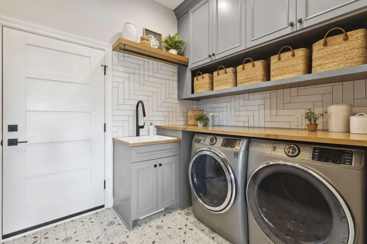 Aménager une buanderie moderne avec lave-linge, sèche-linge, évier gris et paniers en osier sur étagères.