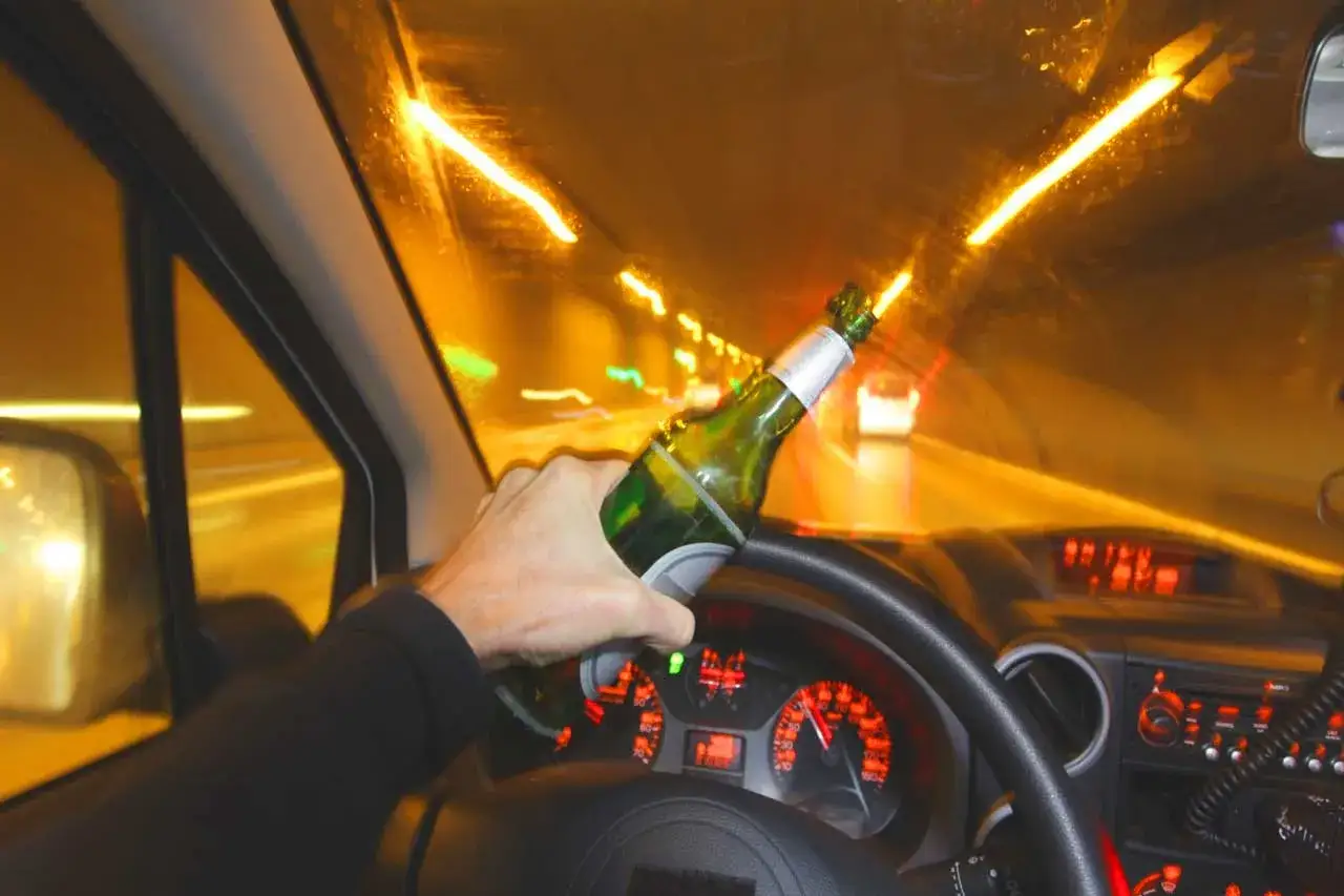 Ręka trzymająca butelkę piwa za kierownicą. Jazda pod wpływem alkoholu to nie dopuszczalna ilość alkoholu i ogromne ryzyko.