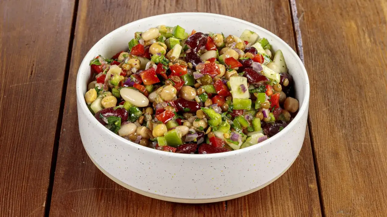 Una ciotola piena di colorata insalata di legumi, con ceci, fagioli, lenticchie, peperoni, cipolla rossa e prezzemolo, su un tavolo di legno.