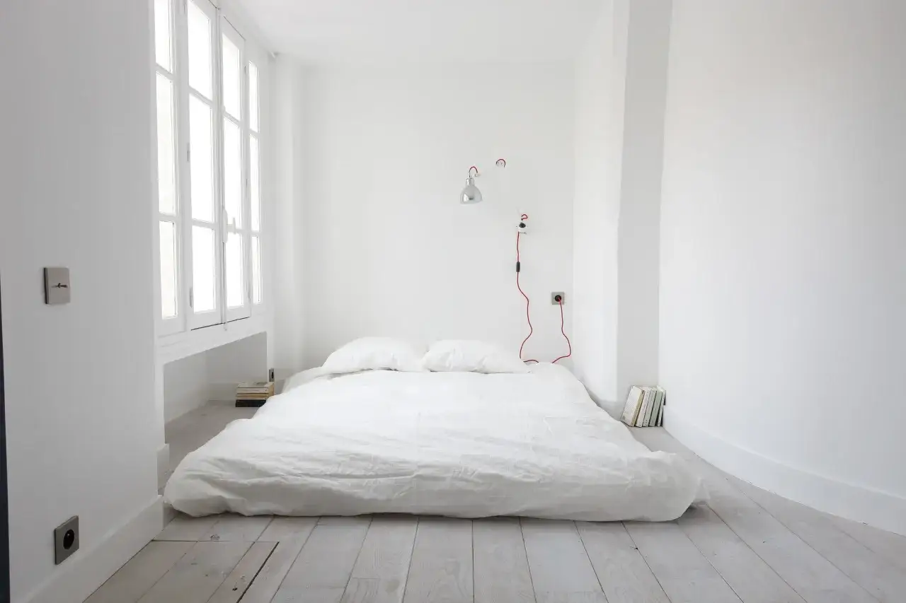 Chambre minimaliste avec un matelas au sol sans sommier, deux oreillers, une lampe murale rouge et des livres empil&eacute;s.