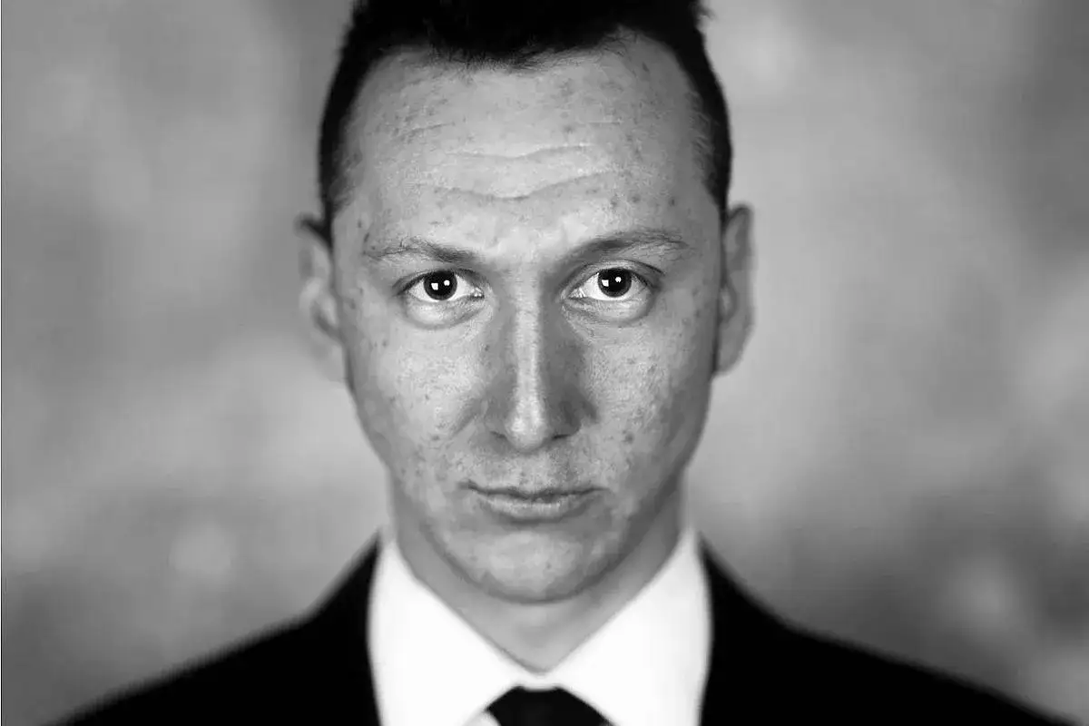 Czarno-białe zdjęcie mężczyzny w garniturze. Jego spojrzenie jest intensywne, a na twarzy widać drobne niedoskonałości. To taki **bodo kox** portret.