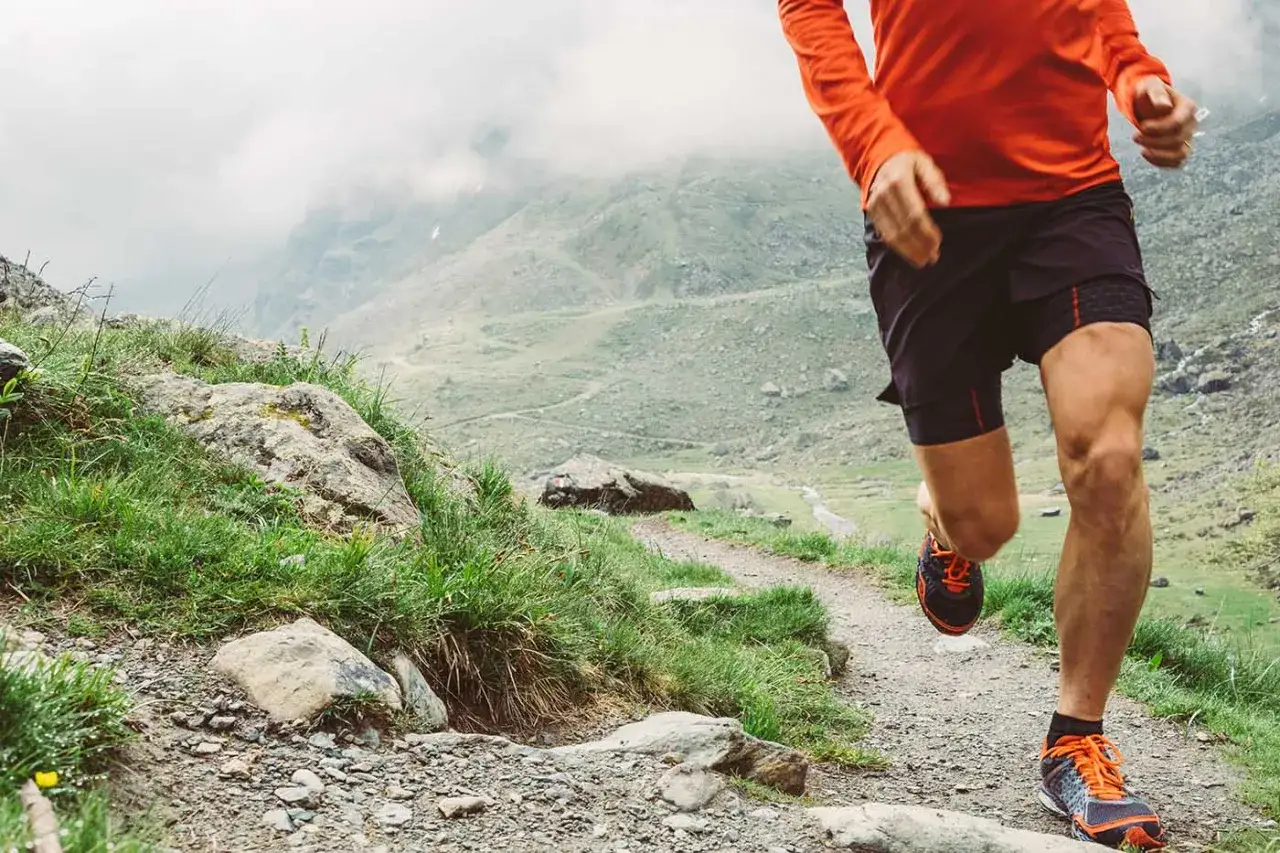 Ein L&auml;ufer im orangen Shirt und schwarzen Shorts absolviert sein trailrunning training auf einem steinigen Bergpfad.