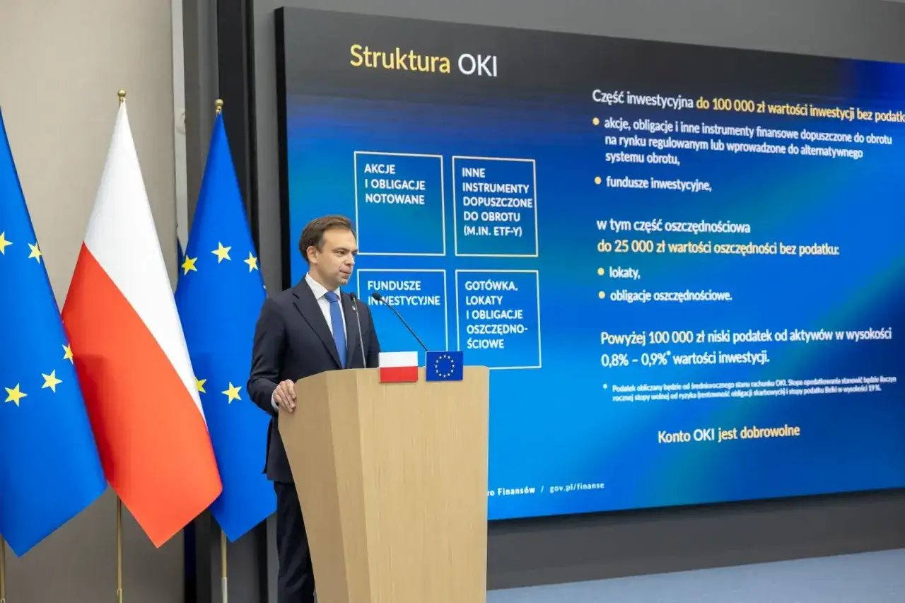Mężczyzna przemawia przy mównicy, obok flagi Polski i UE. Na ekranie struktura OKI, wyjaśniająca zmiany w podatku Belki.