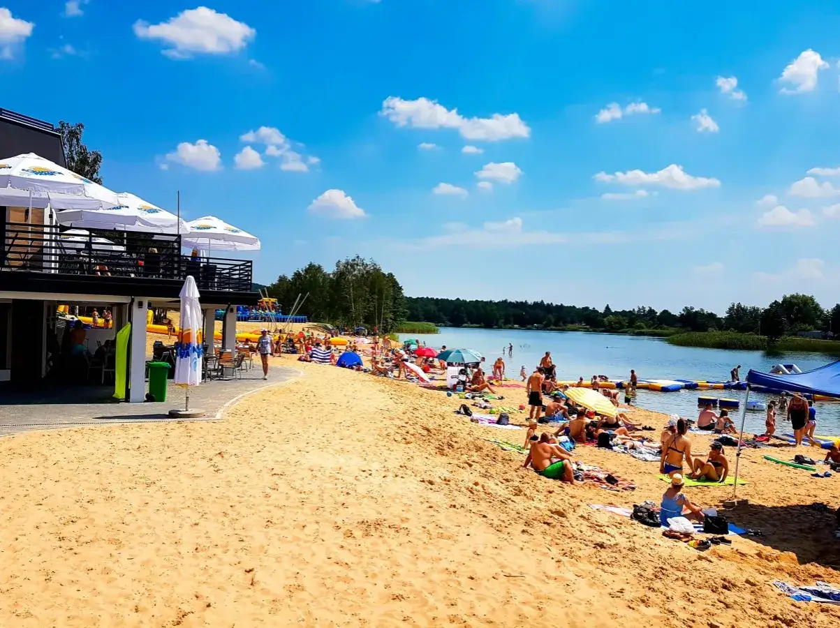 Słoneczny dzień nad zalewem Bolmin. Ludzie odpoczywają na piaszczystej plaży, kąpią się i korzystają z wodnych atrakcji.