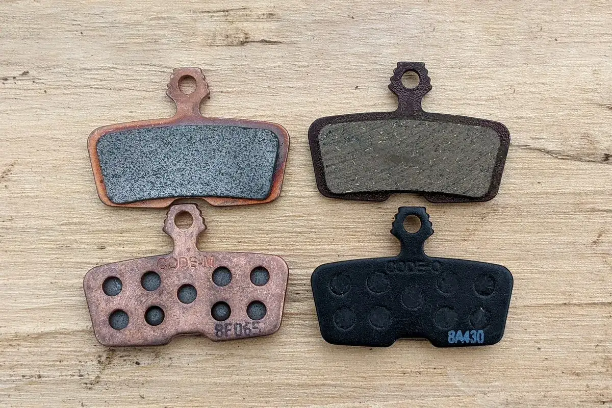 Two pairs of sintered brake pads, one copper and one black, sit on a wooden surface. These durable sintered brake pads offer superior stopping power.