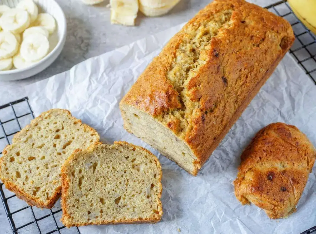 Ein goldbraunes, saftiges Banana Bread liegt auf Backpapier, daneben zwei Scheiben und ein kleines Stück. Bananenscheiben in einer Schüssel.