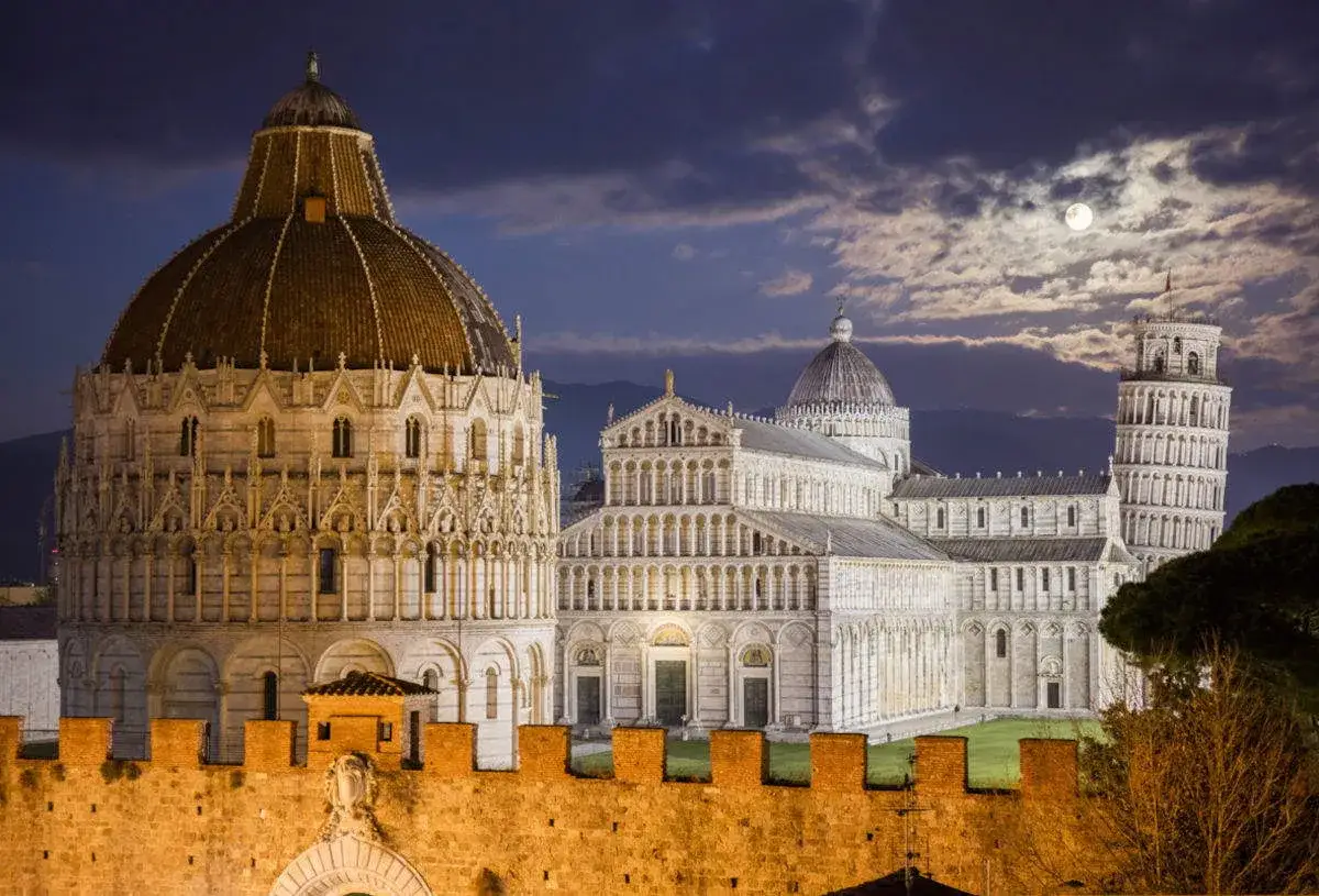 La Piazza dei Miracoli di Pisa, con il Battistero, il Duomo e la Torre Pendente, illuminati dalla luna piena sotto un cielo nuvoloso.