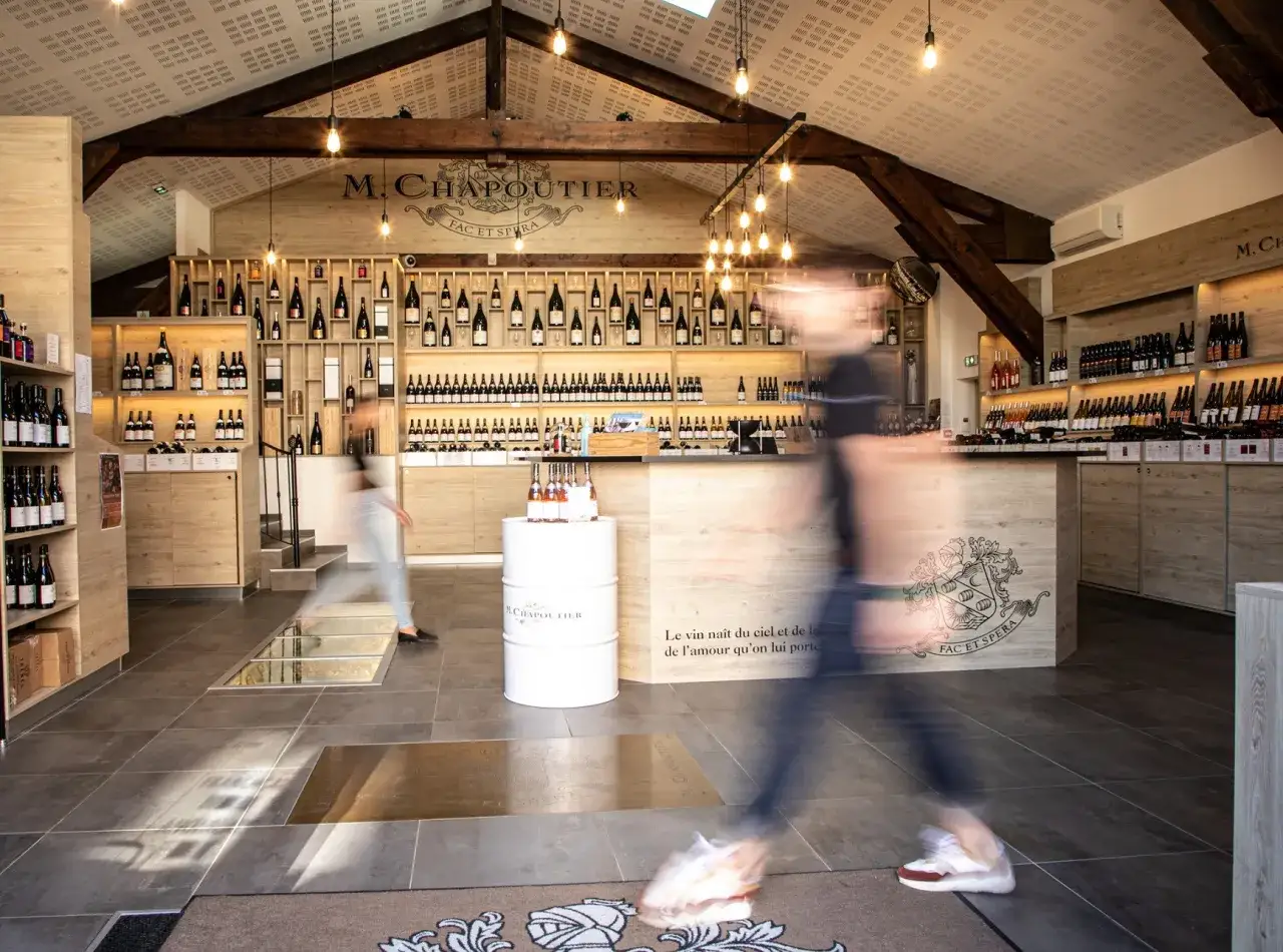 Une **soirée dégustation vin** dans une cave M. Chapoutier. Des étagères remplies de bouteilles, des lumières chaudes et des clients flous en mouvement.