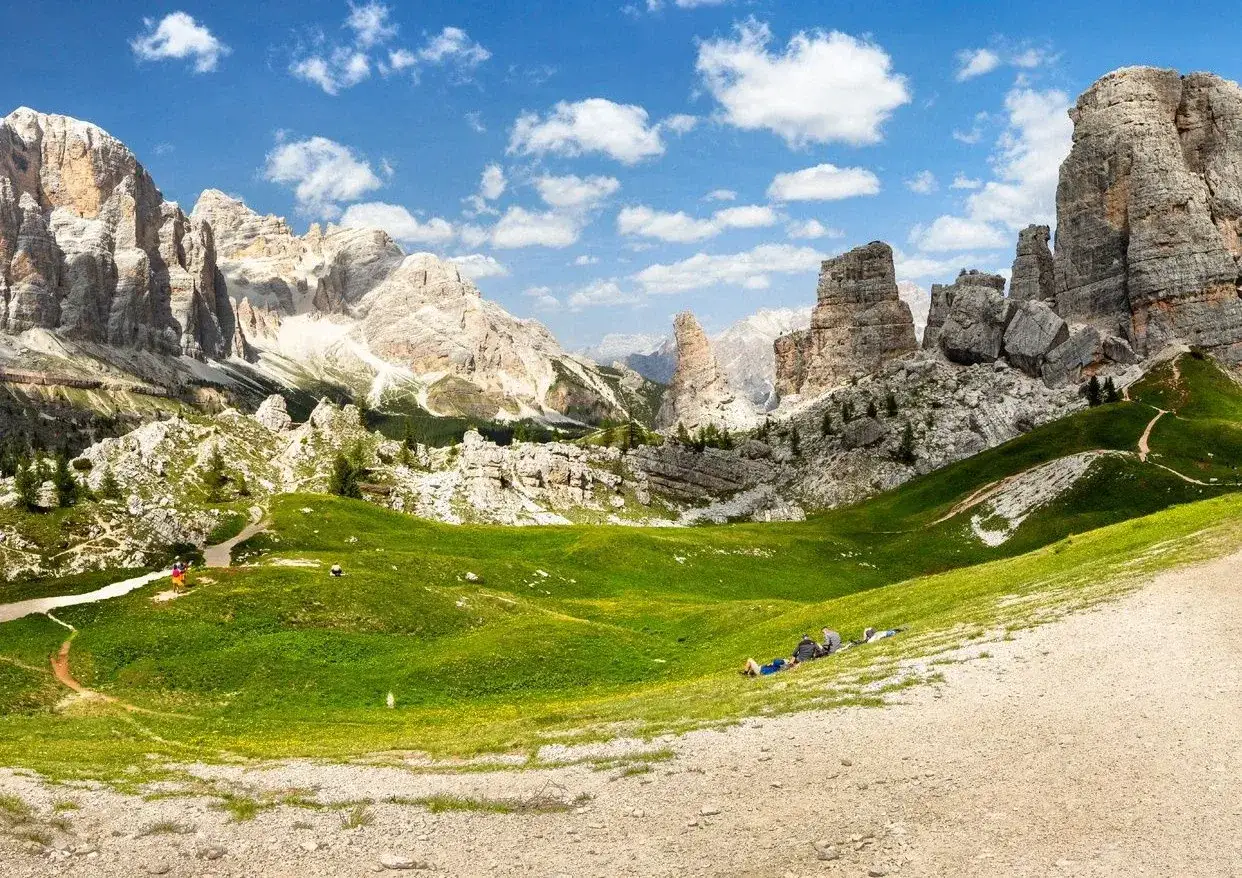 Un paesaggio mozzafiato con le iconiche **5 torri** delle Dolomiti, un prato verdeggiante e un cielo azzurro con nuvole bianche.