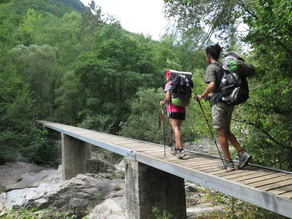 Dos excursionistas con mochilas cruzan un puente de madera en una senda pirenaica.
