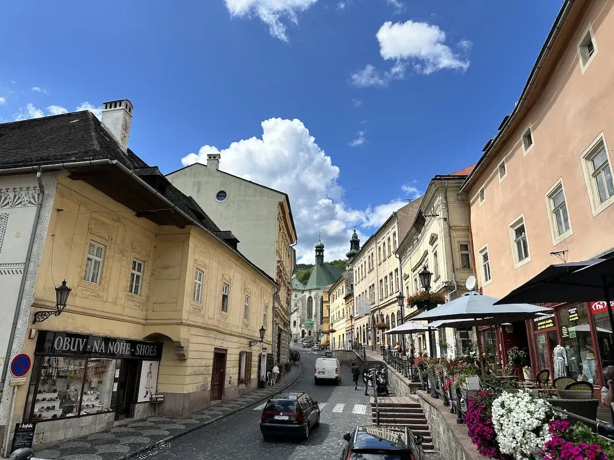 Blick auf eine charmante Straße in Banská Štiavnica mit historischen Gebäuden, Cafés und blühenden Blumen unter blauem Himmel.