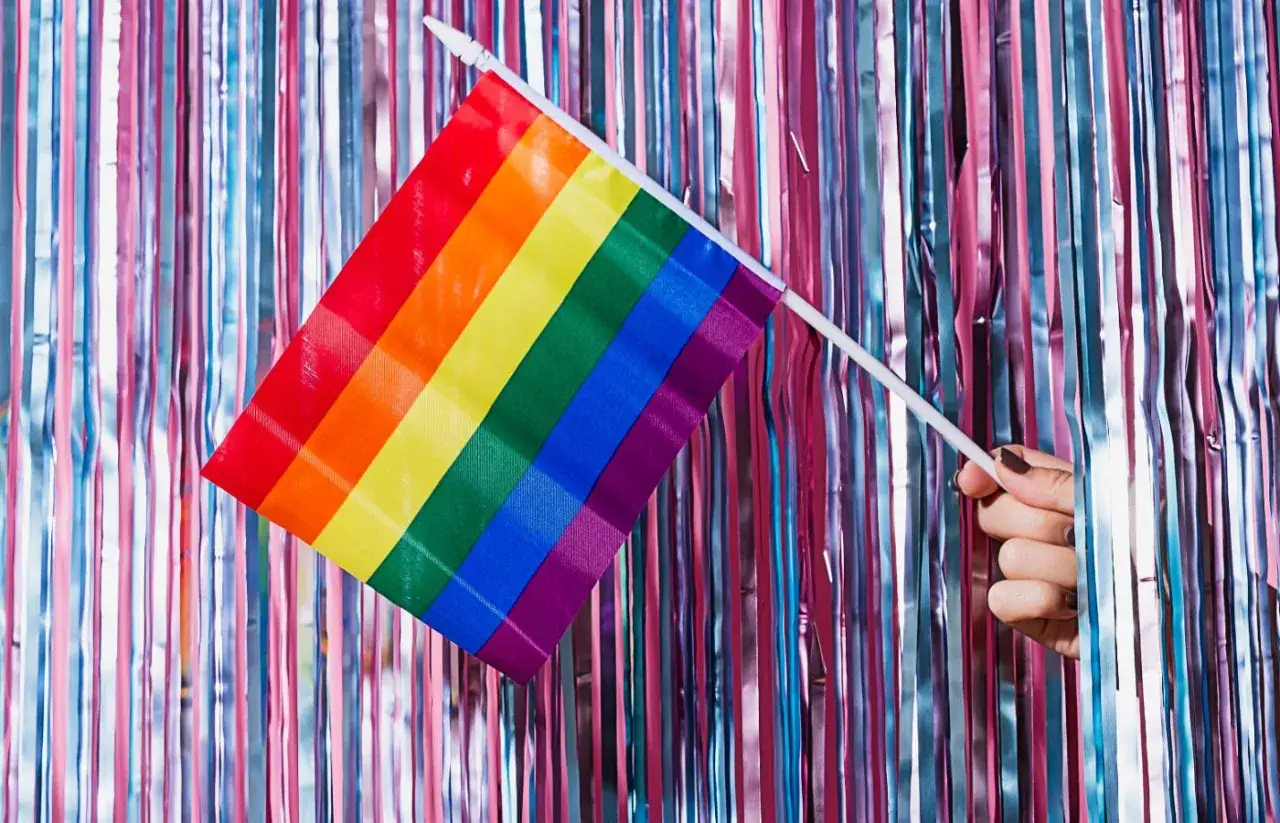 Tęczowa flaga w dłoni na tle błyszczących zasłon. To symbol walki z homofobią i akceptacji.