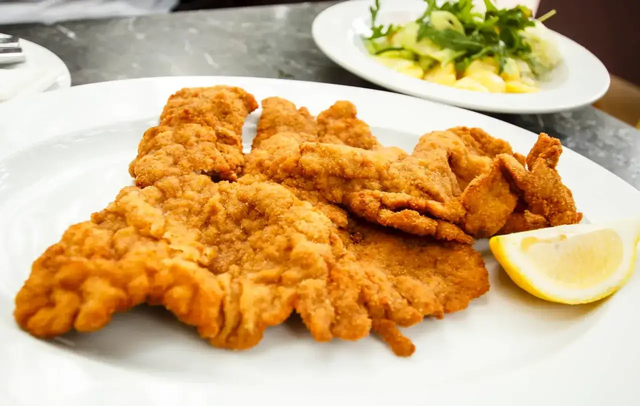 Ein knuspriges Wiener Schnitzel auf einem wei&szlig;en Teller, garniert mit einer Zitronenspalte. Daneben ein kleiner Salat.