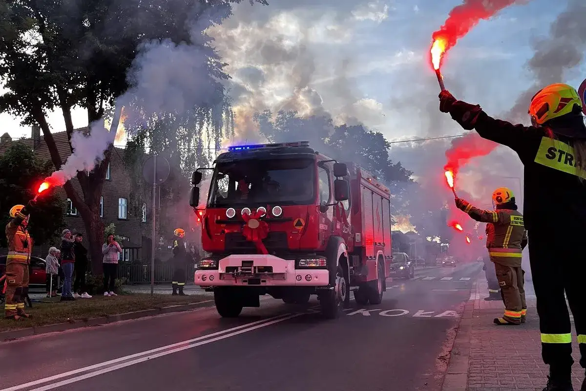 Uroczysta parada strażacka z dymem i flarami. Zabudowa pożarnicza z ozdobną kokardą na masce.