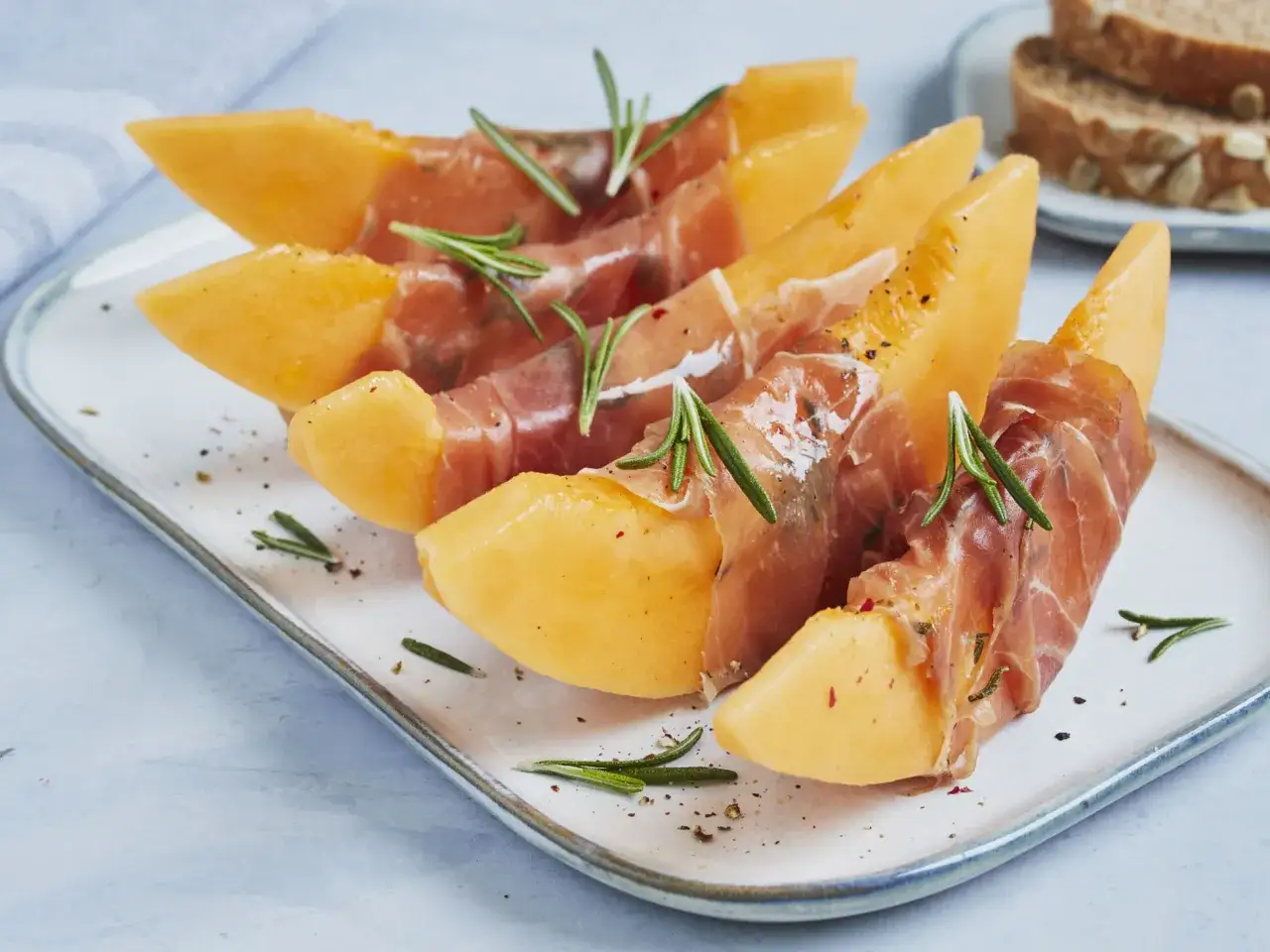 Saftige Honigmelone mit zartem Schinken umwickelt, garniert mit Rosmarin und Pfeffer. Ein köstlicher Genuss!