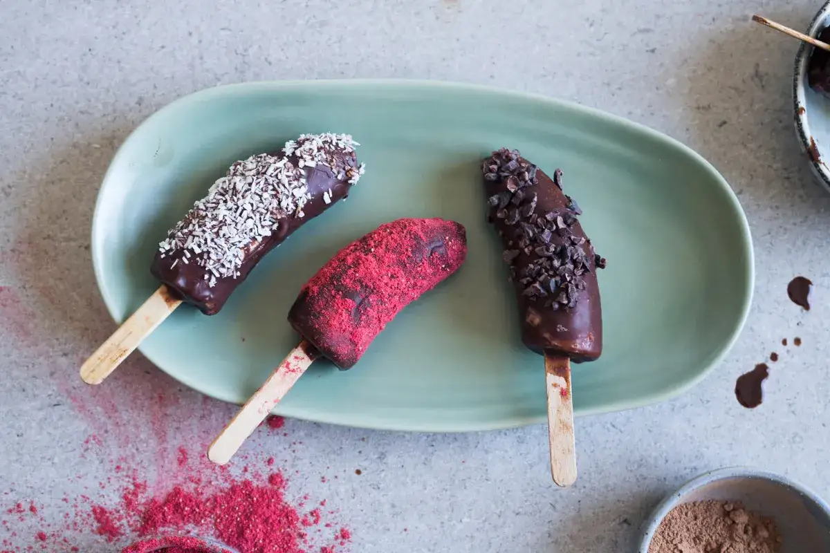 Chokladöverdragna bananer på pinne, toppade med kokos, kakaonibs och hallonpulver. Ett enkelt bananglass recept för en svalkande fika.