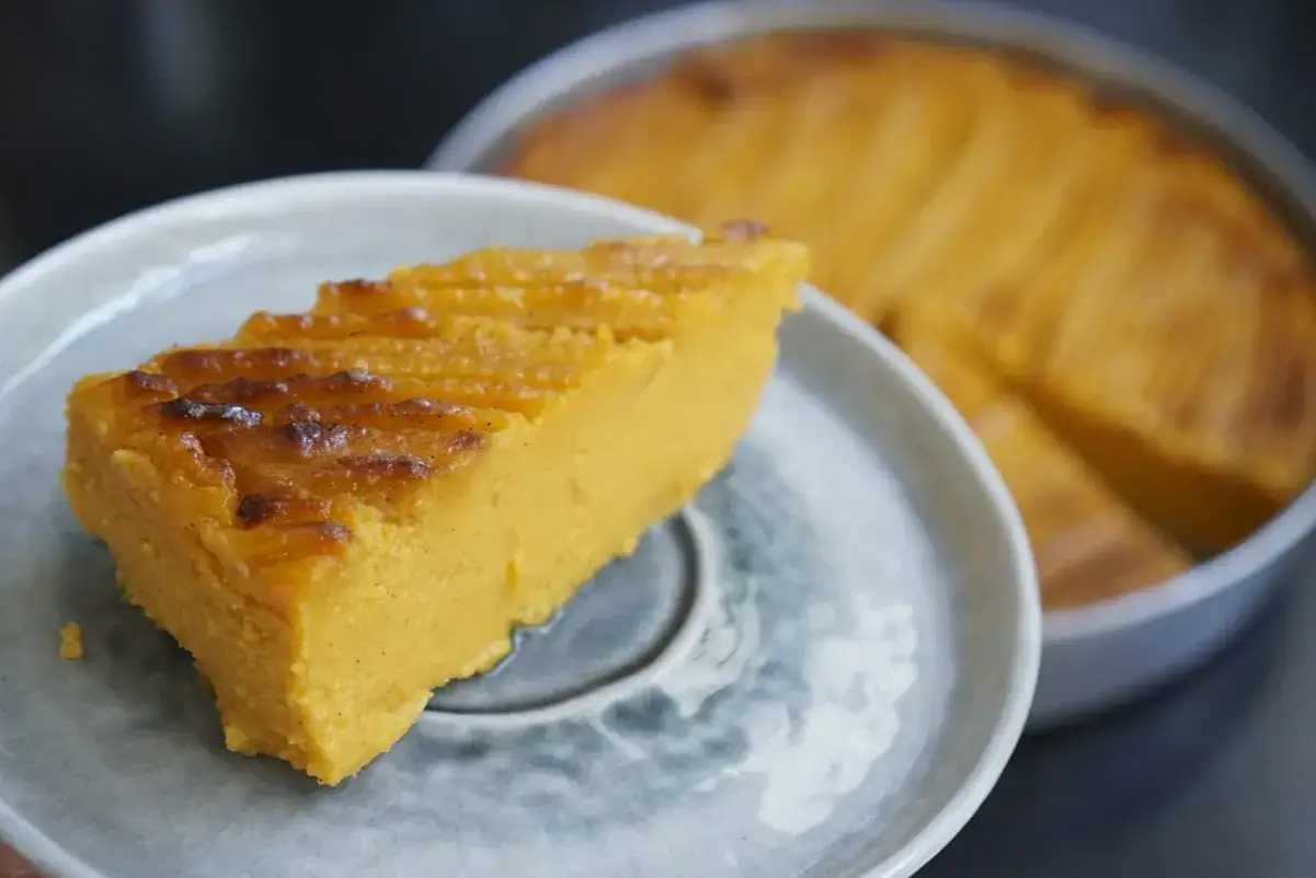 Tranche de g&acirc;teau patate douce, dor&eacute;e et moelleuse, servie sur une assiette grise. Le reste du g&acirc;teau est visible en arri&egrave;re-plan.