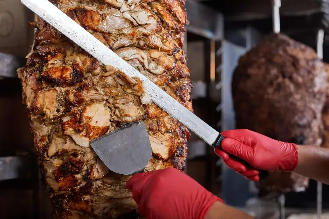 Ręce w czerwonych rękawiczkach kroją soczyste mięso z rożna. Gdzie kupić pyszny mbm kebab?