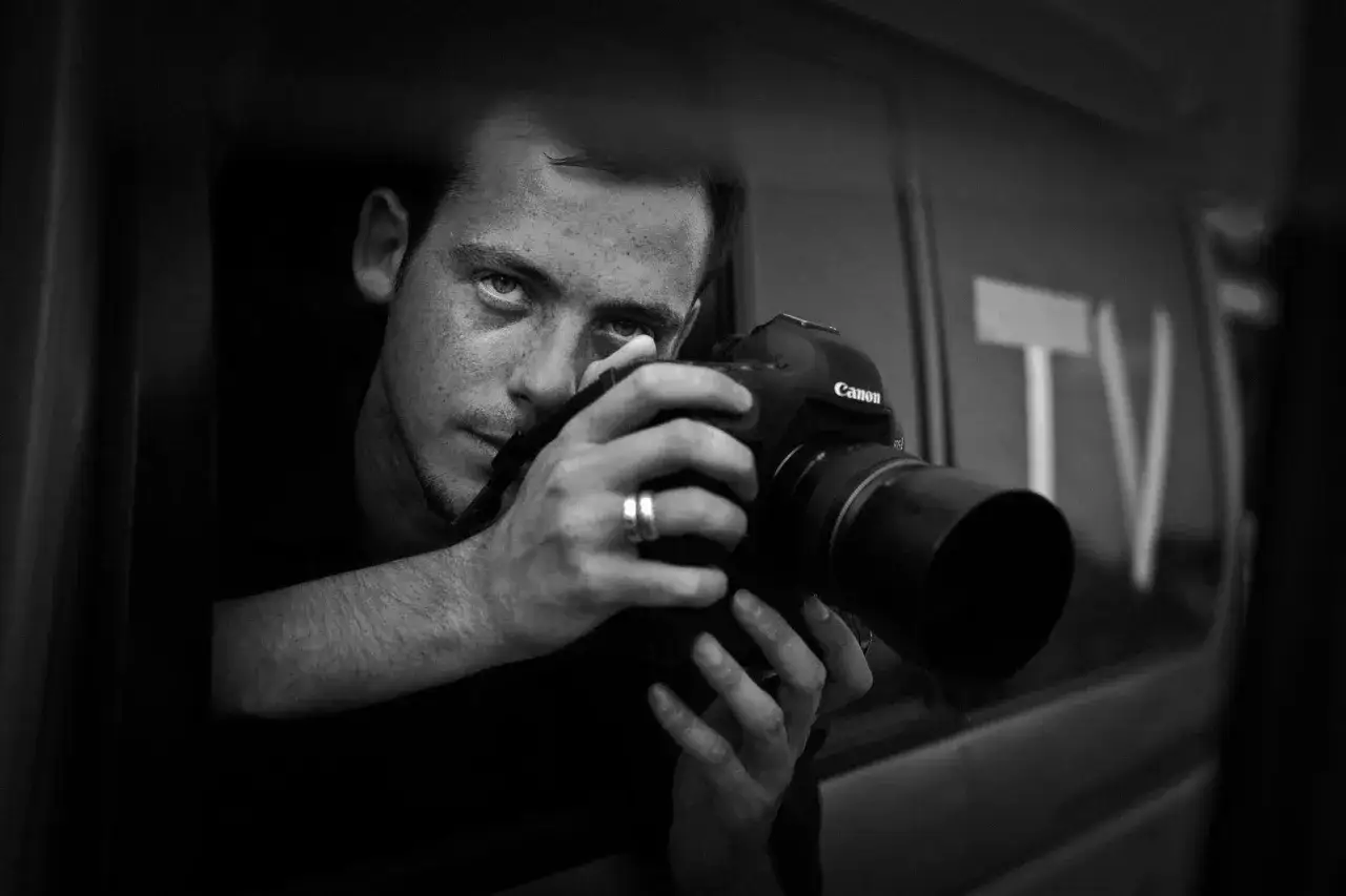 Mężczyzna z aparatem Canon, z pierścionkiem na palcu, spogląda zza okna. To obrazek dla tych, którzy chcą wiedzieć, jak zostać fotoreporterem.
