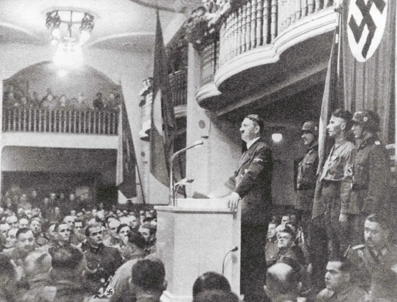 Adolf Hitler spricht vor einer Menge. Hinter ihm stehen Soldaten. Ein Hakenkreuz-Banner ist sichtbar. Dies ist eine Szene, die an das Attentat auf Hitler 1939 erinnert.