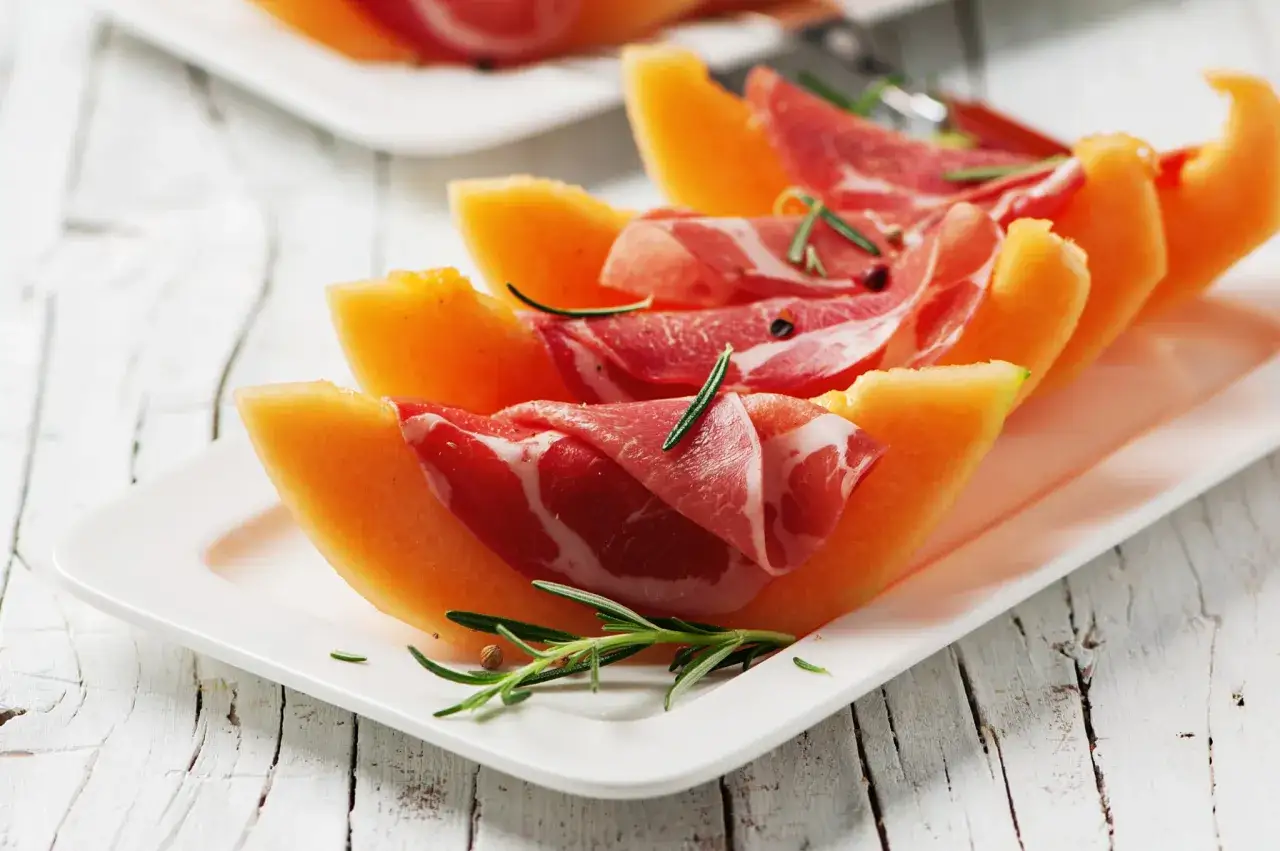 Tranches de melon et jambon cru sur une assiette blanche, garnies de romarin. Une id&eacute;e parfaite pour savoir quel vin avec melon et jambon cru.