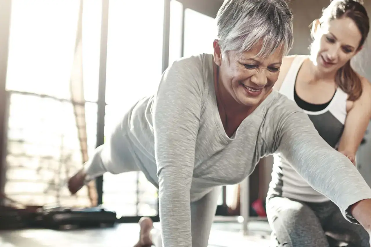 Eine Frau mit grauen Haaren lächelt, während sie mit Unterstützung einer Trainerin eine Yoga-Pose macht. Nach Krebserkrankung nicht mehr belastbar, findet sie neue Kraft.