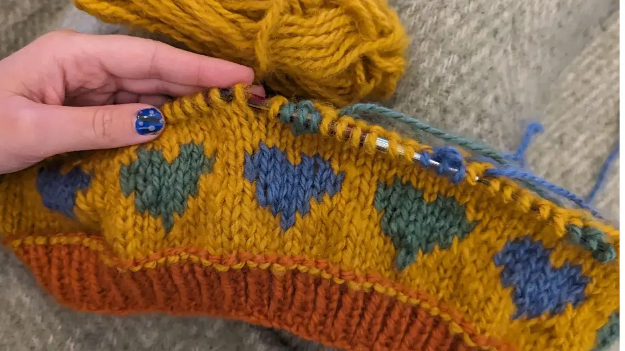 Main tricot jacquard avec des c&oelig;urs bleus et verts sur fond jaune moutarde. Une main avec un ongle d&eacute;cor&eacute; tient les aiguilles.