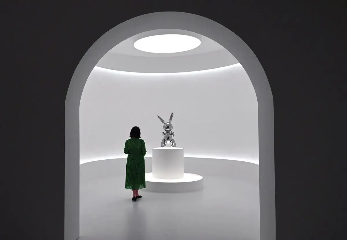 A woman in a green dress admires a shiny balloon rabbit sculpture, a prime example of blue chip art, in a minimalist gallery.