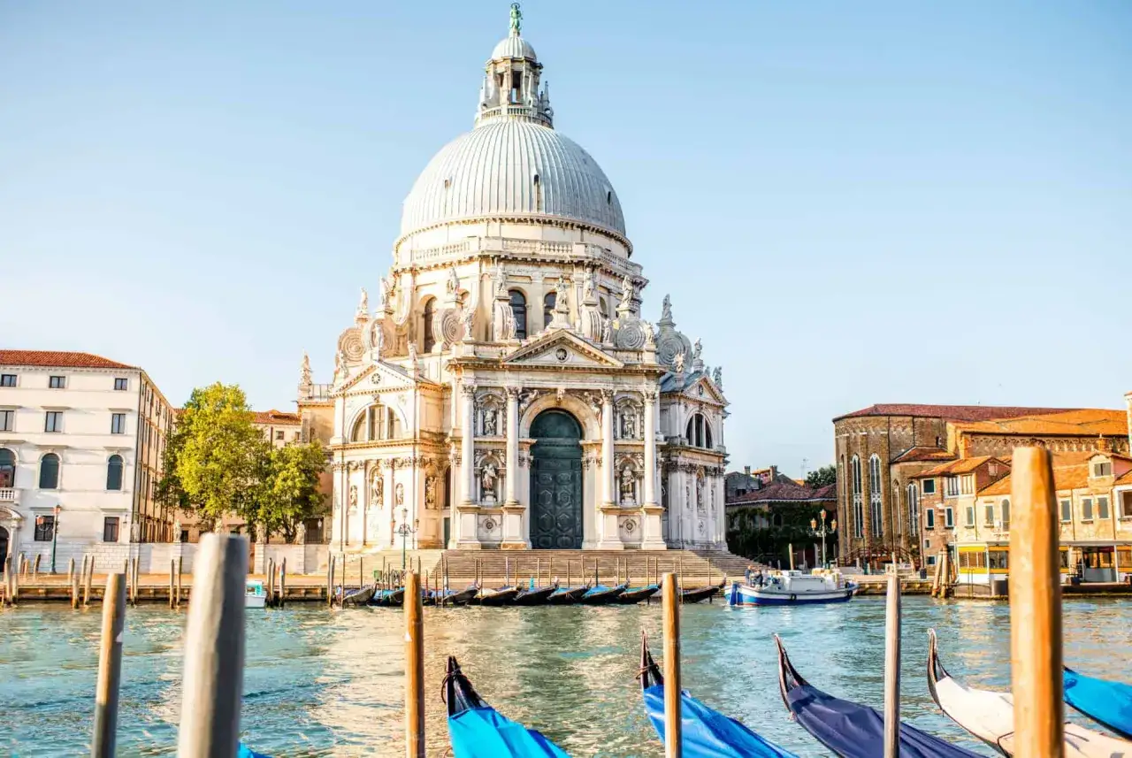 La basilique Santa Maria della Salute, majestueuse, se dresse à Venise, ses dômes blancs reflétant le ciel bleu. Des gondoles bleues et noires attendent sur le Grand Canal.