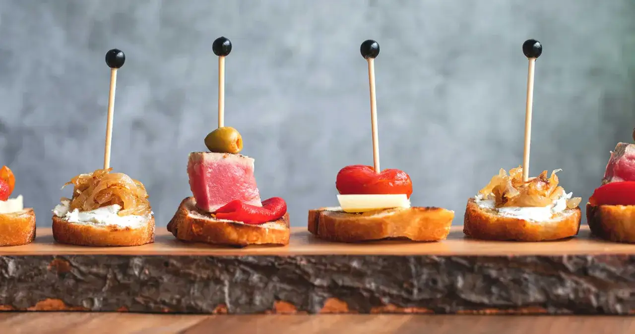 A row of appetizers on a wooden board, including a smoked trout dip with caramelized onions, tuna with olives, and tomato with cheese.