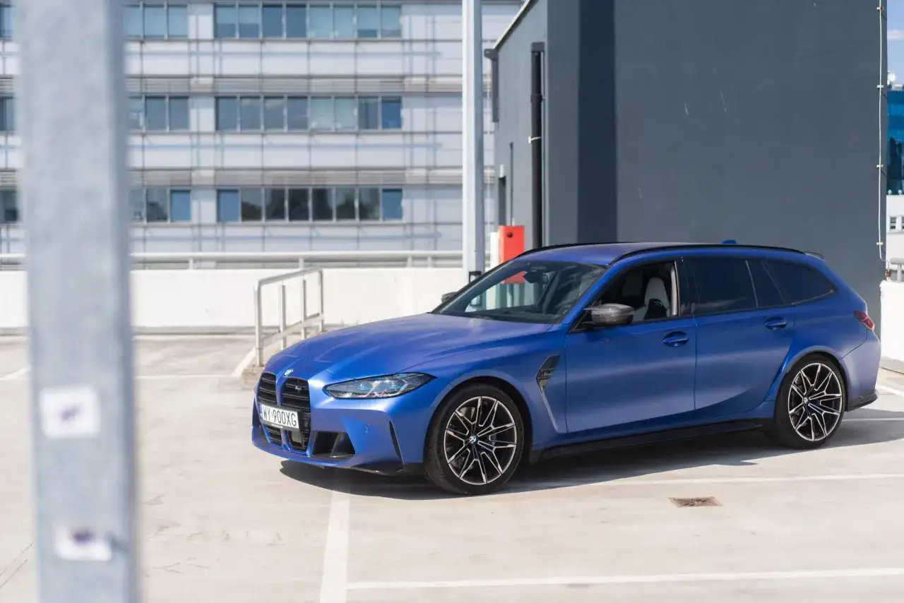 Niebieskie BMW M3 Touring na parkingu, z nowoczesnym budynkiem w tle.