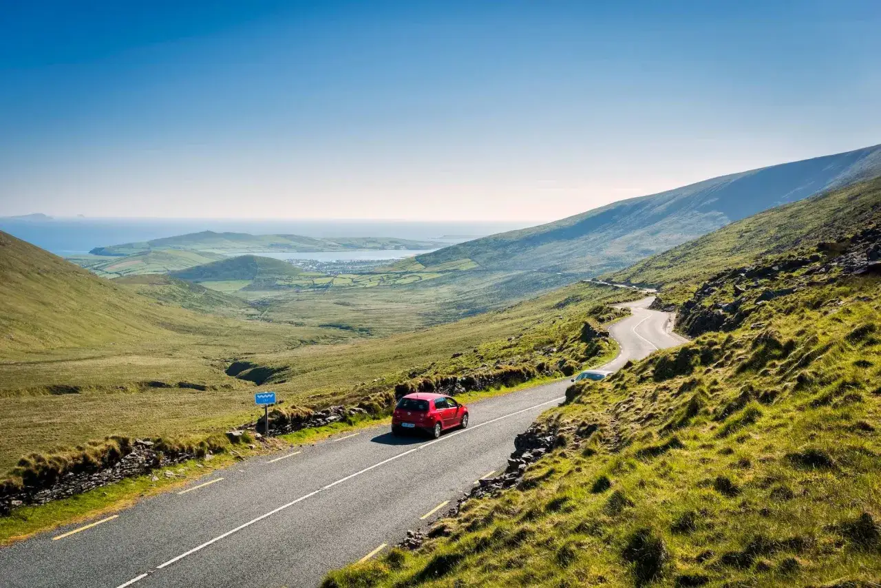 A red car drives on a winding road through green Irish hills, offering a scenic view perfect for an Ireland itinerary.