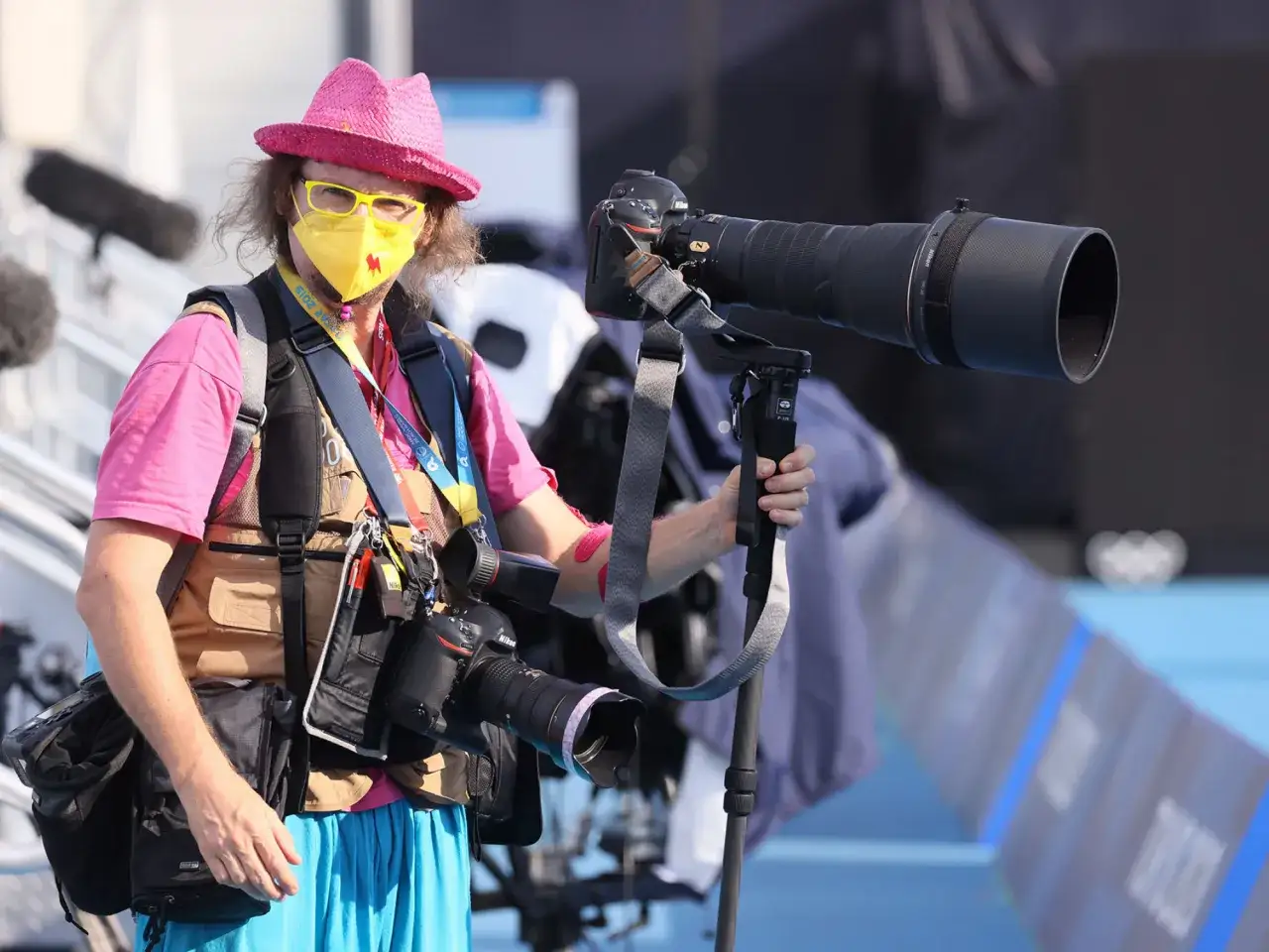 Adam Nurkiewicz, fotograf w różowej czapce i żółtej maseczce, z dwoma aparatami i długim obiektywem, gotowy do uchwycenia sportowych emocji.