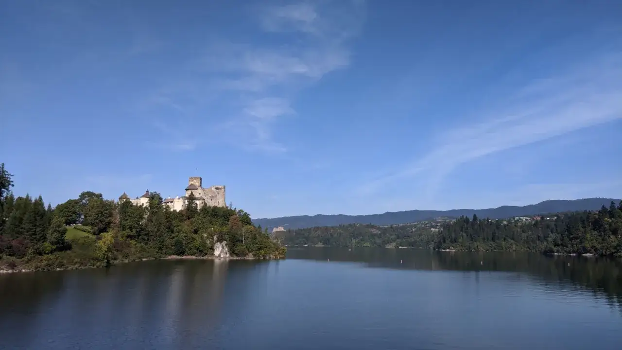 Zamek Czorsztyn góruje nad jeziorem, otoczony zielenią. W oddali widać kolejne ruiny i zalesione wzgórza.