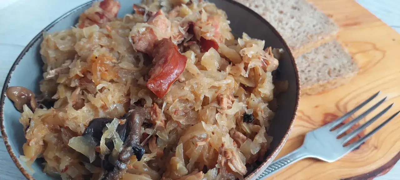 Pyszny bigos, który Ania gotuje, w misce z widocznymi kawałkami mięsa i grzybów. Obok kromki chleba i widelec.