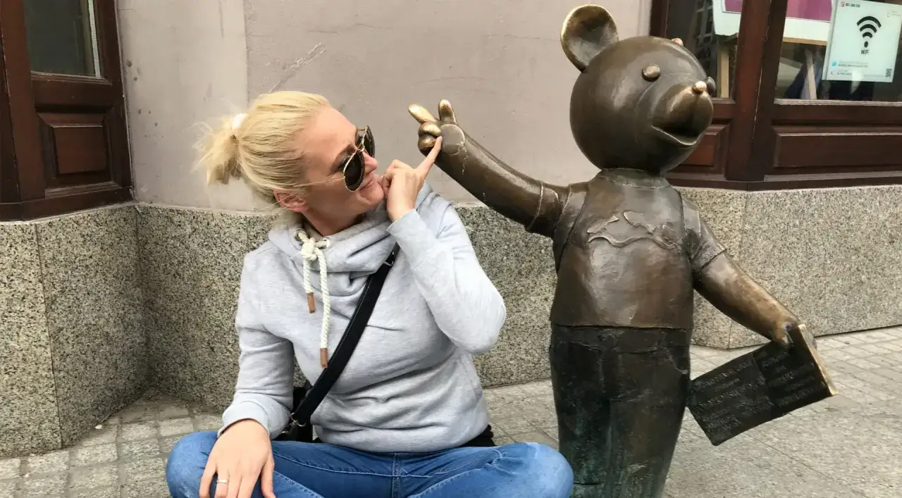 Kobieta w okularach przeciwsłonecznych i szarej bluzie bawi się z pomnikiem misia, który trzyma tabliczkę. To część szlaku łodzi bajkowej.