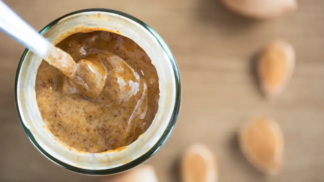 Une cuillère plonge dans un pot de purée d'amande maison, crémeuse et onctueuse. Quelques amandes sont disposées autour.