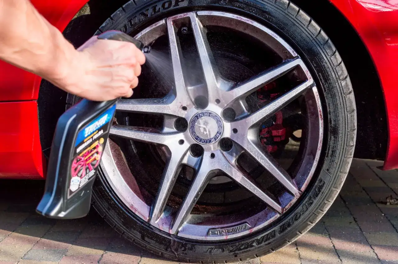 Czyszczenie felg aluminiowych. Ręka z butelką preparatu spryskuje brudną, srebrną felgę Mercedesa.